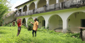 Picture of artist Sharon Pérez in a green landscape with a colonial building, talking to a member of the community