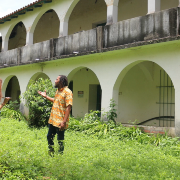 Picture of artist Sharon Pérez in a green landscape with a colonial buildingtalking to