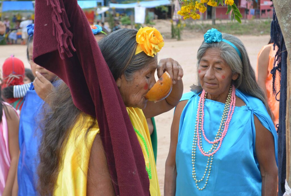 Photo fo two elder women performing a ceremony. One, dressed with yellow colours is drinking from a bowl while holding a maroon flag, while the other, dressed in blue, looks at her